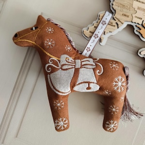 Puede incluir: Adorno de caballo de fieltro marrón con copos de nieve y campanas bordados en blanco. El caballo tiene una cinta blanca para colgar y una melena trenzada. Es una decoración festiva.