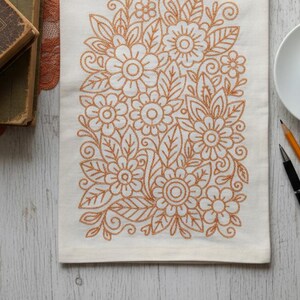 May include: Off-white linen tea towel with an embroidered floral design in shades of orange. The design features flowers, leaves, and swirling vines. The towel is rectangular and lies on a light-colored wooden surface.