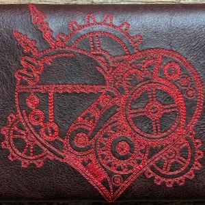 Può includere: Un portafoglio in pelle marrone scuro con un design a cuore steampunk rosso ricamato. Il cuore è composto da ingranaggi, ruote dentate e altri elementi meccanici, creando un motivo unico e intricato. Il portafoglio ha una superficie strutturata.