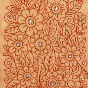 Könnte beinhalten: Nahaufnahme eines pfirsichfarbenen Stoffes mit einem gestickten Blumenmuster. Die Stickerei ist in einem warmen Orangeton gehalten und zeigt Blumen, Blätter und wirbelnde Details. Jede Blumenmitte hat einen kleinen, funkelnden Strassstein.