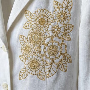 Puede incluir: Blazer blanco con bordado floral dorado en la parte delantera. El diseño floral incluye flores y hojas detalladas, creando un acento decorativo. El blazer tiene solapas clásicas y dos botones blancos.