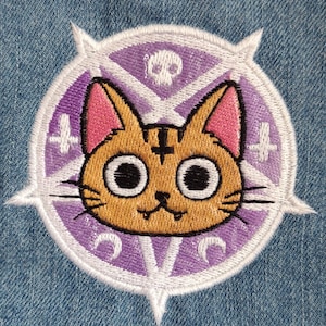 May include: An embroidered patch featuring a cartoon cat with a pentagram on its forehead, surrounded by a purple pentagram with white accents. The cat has large eyes and a mischievous grin.