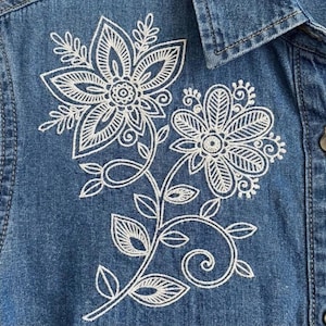 May include: Close-up of a blue denim shirt with white floral embroidery. The embroidery shows two large flowers with detailed petals and leaves. The shirt has a button-down front and a collar.