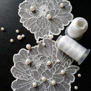 Puede incluir: Dos posavasos de encaje blanco con forma floral, adornados con perlas y dos carretes de hilo blanco sobre una superficie negra. Los posavasos tienen diseños intrincados y detallados. Las perlas dispersas añaden un toque de elegancia.