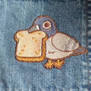 May include: Embroidered patch on denim fabric featuring a gray pigeon holding a brown and yellow slice of bread.