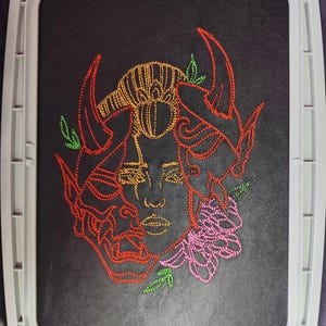 May include: Embroidered design of a face with horns and flowers. The face is outlined in orange, the horns are red, and the flowers are pink and green.