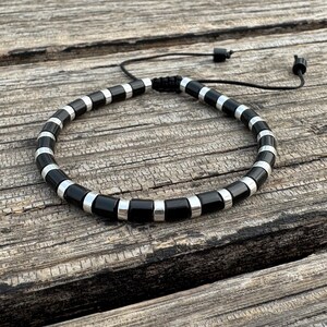 May include: Black and silver beaded bracelet. The bracelet features alternating black rectangular beads and silver cylindrical beads. The adjustable black cord allows for a comfortable fit. The bracelet is displayed on a weathered wooden surface.