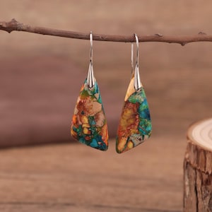 May include: A pair of dangle earrings with a silver-tone metal hook and a colorful, triangular stone pendant. The stone features a mix of turquoise, orange, and brown hues. The earrings are displayed on a brown twig against a blurred wooden background.