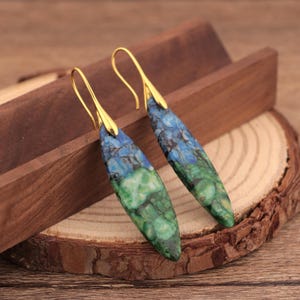 May include: A pair of dangle earrings with a gold-colored hook and a teardrop-shaped stone. The stone features a blend of blue and green hues, creating a marbled effect. The earrings are displayed on a wooden surface.