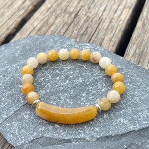 Op de afbeelding: Een armband met gele en crèmekleurige ronde kralen. Een gebogen, rechthoekige gele steen is gecentreerd op de armband, met zilverkleurige metalen accenten. De armband wordt gepresenteerd op een grijze stenen ondergrond.