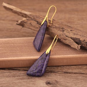 May include: A pair of dangle earrings featuring elongated, triangular purple stones with gold-colored hooks. The earrings are displayed on a wooden surface with a piece of driftwood.