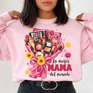 May include: Pink sweatshirt featuring a bouquet of makeup products, including eyeshadow, lipstick, and brushes. The bouquet is tied with a pink ribbon and adorned with yellow flowers and the text "La mejor MAMA del mundo" in red.