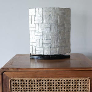 Puede incluir: Una lámpara plateada con una pantalla rectangular con diseño de mosaico. La pantalla está hecha de pequeñas piezas rectangulares dispuestas en un patrón de ladrillos. La lámpara está sobre una base negra y sobre una mesa de madera.