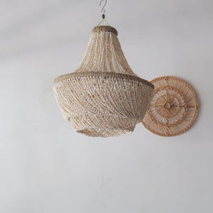 May include: A large, tiered chandelier with a beige and white beaded design. The chandelier is suspended from a hook and features a woven, light brown trim. A circular woven wall hanging is to the right.
