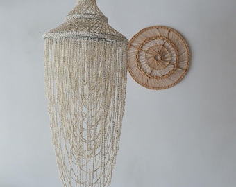 May include: A large, cream-colored seashell chandelier with a conical shape and cascading strands of shells. A round, woven wall hanging is to the right. The chandelier is suspended from the ceiling, creating a coastal or bohemian aesthetic.
