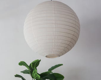 May include: A round, off-white paper lantern hanging from a black cord. The lantern has horizontal lines and a circular shape. Green leafy plant in the foreground. The lantern is a decorative lighting fixture.