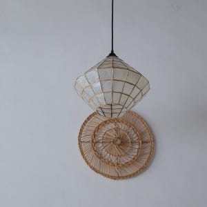 May include: A hanging pendant light with a geometric, diamond-shaped shade made of white panels and gold-coloured metal framing. Below it is a round, woven wall hanging made of natural-coloured fibres. The light hangs from a black cord.