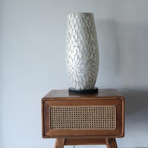 Op de afbeelding: Een hoge, parelmoerwitte lamp met een bloemmotief staat op een donkere basis. De lamp staat op een houten nachtkastje met een geweven rotan paneel op de lade. Het nachtkastje heeft een modern design.