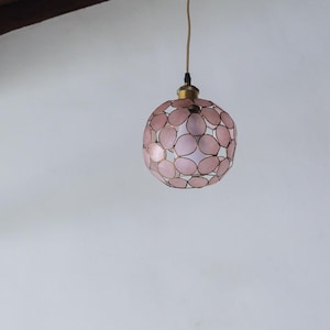 May include: A spherical pendant light fixture with a pink, petal-like design. The light has a brass-colored top and a white interior, suspended from a thin, gold-colored cord. The fixture is hanging against a white background.
