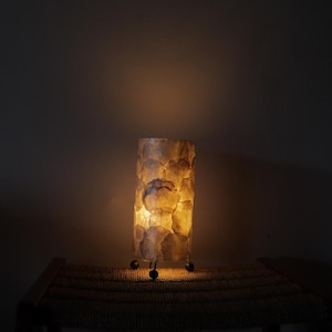 May include: A cylindrical lamp with a textured, off-white shade. The lamp is illuminated from within, casting a warm glow. It stands on a small, dark-colored base with three small feet. The lamp is placed on a woven surface.