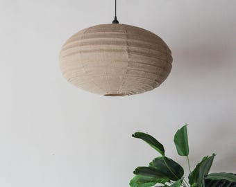 May include: A beige, spherical pendant light fixture with a textured, horizontal-lined design. The light hangs from a black cord and is suspended against a white wall. A green plant is visible in the lower right corner.