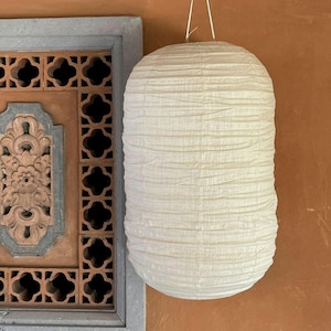 May include: A cream-coloured, cylindrical paper lantern hangs against a warm, orange-brown wall. The lantern has a textured, pleated surface and is suspended by thin white strings. A decorative wooden window frame with carved details is partially visible.