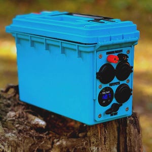 May include: A blue portable power station with multiple outlets and a digital display showing the voltage. The power station is sitting on a tree stump.