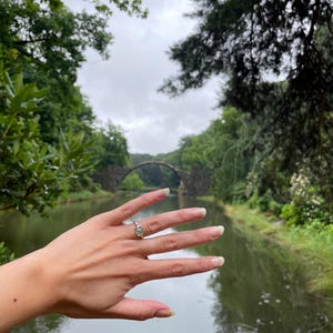 Pu&ograve; includere: Una mano che indossa un anello d'argento con una pietra preziosa verde chiaro, sullo sfondo di un ponte di pietra su un corso d'acqua. La scena &egrave; circondata da lussureggianti alberi verdi sotto un cielo nuvoloso.