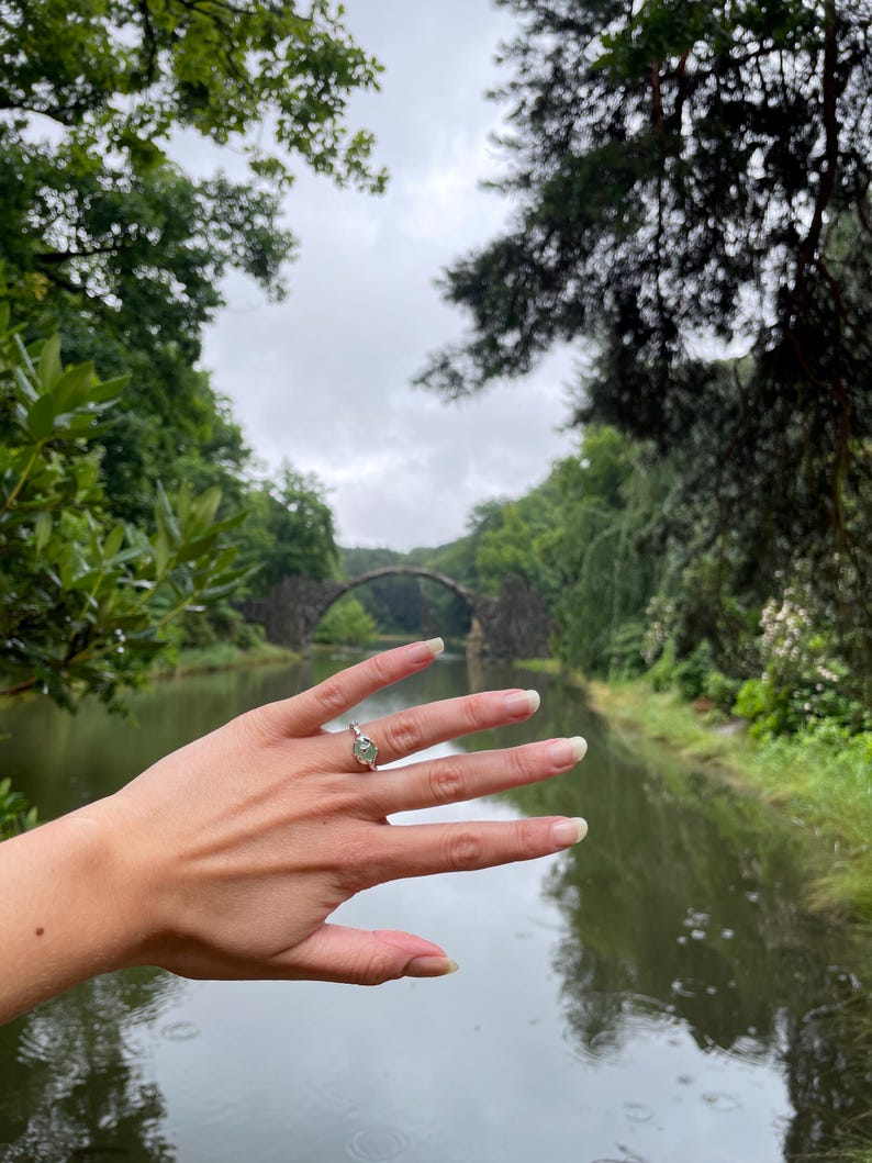 Pu&ograve; includere: Una mano che indossa un anello d'argento con una pietra preziosa verde chiaro, sullo sfondo di un ponte di pietra su un corso d'acqua. La scena &egrave; circondata da lussureggianti alberi verdi sotto un cielo nuvoloso.