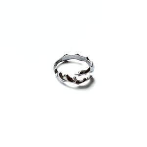 May include: A silver-colored ring with an organic, flowing design. The ring has an irregular, textured surface with a wave-like pattern. The ring is set against a white background, highlighting its metallic sheen. The ring is a piece of jewelry.