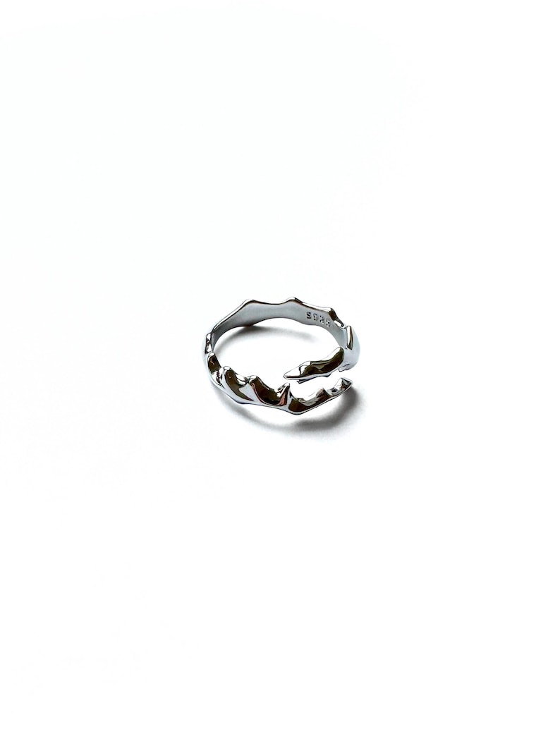 May include: A silver-colored ring with an organic, flowing design. The ring has an irregular, textured surface with a wave-like pattern. The ring is set against a white background, highlighting its metallic sheen. The ring is a piece of jewelry.