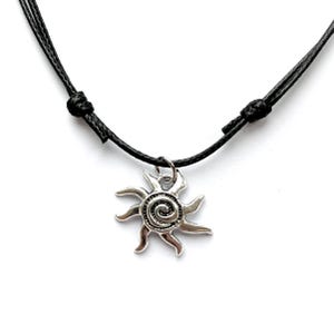 May include: A silver sun pendant necklace featuring a spiral design and spiky rays. The pendant is suspended from a black cord necklace with adjustable knots. The necklace is set against a white backdrop.