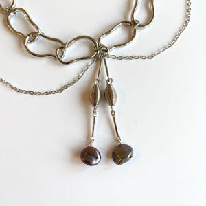 Op de afbeelding: Een zilverkleurige ketting met een abstract kettingontwerp. Twee strengen kleinere kettingen hangen aan de hoofdketting, elk met een zilveren spiraal en een donkere, peervormige kraal. De ketting wordt gepresenteerd op een wit oppervlak.