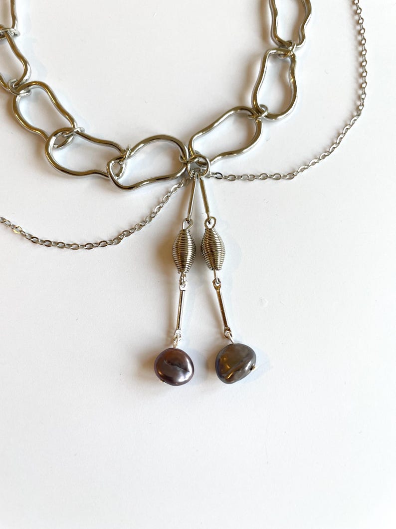 Op de afbeelding: Een zilverkleurige ketting met een abstract kettingontwerp. Twee strengen kleinere kettingen hangen aan de hoofdketting, elk met een zilveren spiraal en een donkere, peervormige kraal. De ketting wordt gepresenteerd op een wit oppervlak.