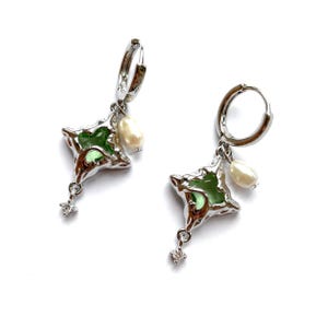 Può includere: Un paio di orecchini a cerchio argentati. Ogni orecchino presenta un ciondolo verde a forma di stella, una perla e un piccolo cristallo pendente. Sfondo bianco.