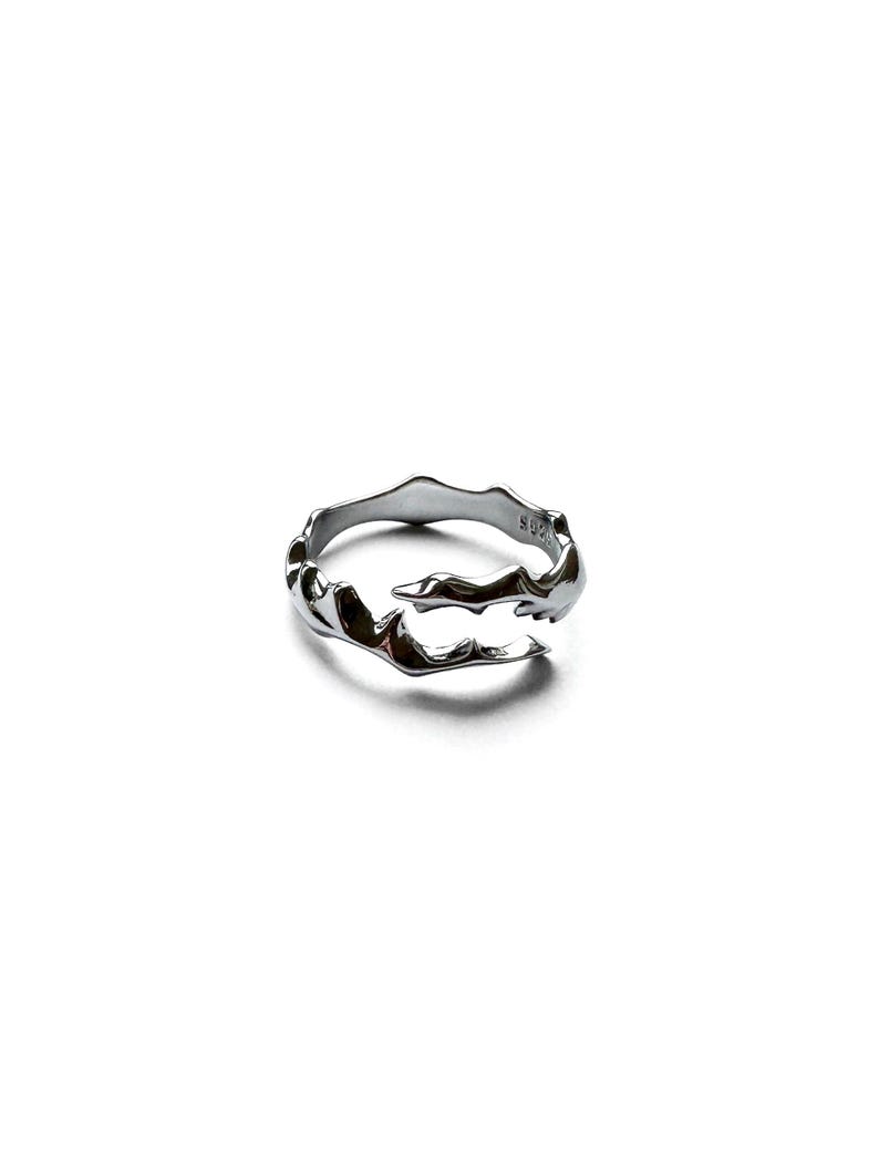 May include: A silver-colored ring with an organic, flowing design. The ring has an irregular, textured surface, with a wave-like pattern. The ring is made of a shiny, metallic material and is set against a white background.