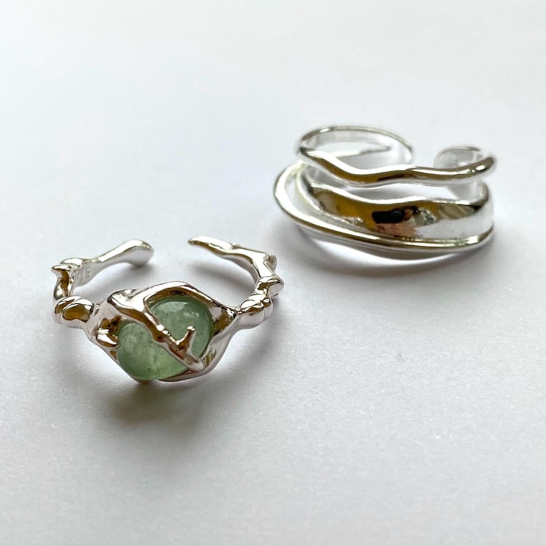 Pu&ograve; includere: Due anelli d'argento. Uno presenta una pietra preziosa verde chiaro incastonata in una fascia strutturata in stile organico. Il secondo anello ha un design a strati e fluido. Entrambi gli anelli sono su una superficie bianca.