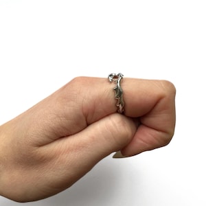 May include: Silver-toned ring with a unique, spiky design. The ring is worn on a finger, showcasing its intricate details and metallic sheen. The ring's design features a series of pointed elements, creating a textured and eye-catching appearance.