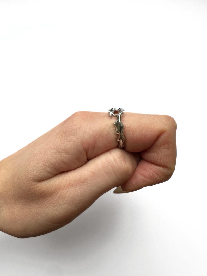 May include: Silver-toned ring with a unique, spiky design. The ring is worn on a finger, showcasing its intricate details and metallic sheen. The ring's design features a series of pointed elements, creating a textured and eye-catching appearance.