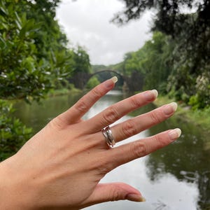 Pu&ograve; includere: Un anello d'argento dal design ondulato &egrave; indossato su una mano. L'anello ha una finitura lucida e un'estetica moderna. Lo sfondo presenta una vista sfocata di un ponte sull'acqua.