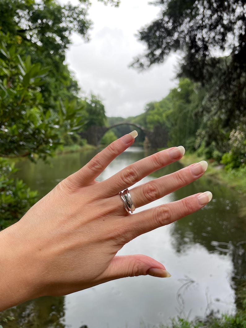 Pu&ograve; includere: Un anello d'argento dal design ondulato &egrave; indossato su una mano. L'anello ha una finitura lucida e un'estetica moderna. Lo sfondo presenta una vista sfocata di un ponte sull'acqua.