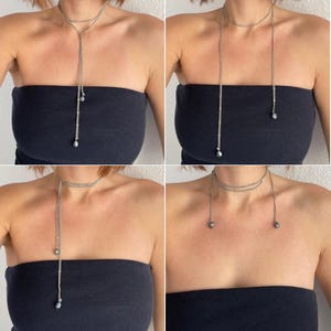 Puede incluir: Collar lariat plateado con una cadena delicada y pequeños detalles de perlas gris oscuro. El collar se muestra en múltiples variaciones, drapeado sobre una persona que lleva un top negro sin tirantes. El collar tiene un estilo gargantilla en capas.