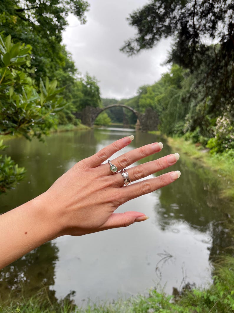 Pu&ograve; includere: Una mano che indossa tre anelli, tra cui un anello di fidanzamento con una pietra preziosa verde chiaro. Gli anelli sono color argento. Lo sfondo presenta un ponte di pietra su un corso d'acqua, circondato da una vegetazione lussureggiante sotto un cielo nuvoloso.