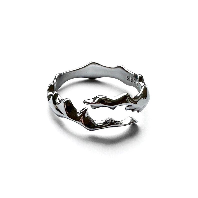 May include: A silver-colored ring with an organic, flowing design. The ring has an open, adjustable style with a textured surface. The metal ring is on a white background. The ring is marked with "S925".