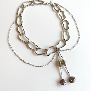 Op de afbeelding: Een zilverkleurige ketting met een open schakelkettingontwerp. De ketting heeft een delicate ketting en twee bungelende strengen met decoratieve kralen en donkergrijze, druppelvormige hangers. De sieraden worden op een wit oppervlak weergegeven.