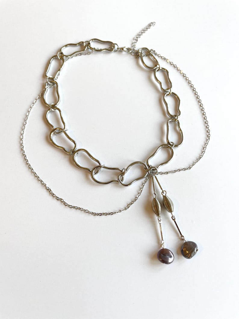 Op de afbeelding: Een zilverkleurige ketting met een open schakelkettingontwerp. De ketting heeft een delicate ketting en twee bungelende strengen met decoratieve kralen en donkergrijze, druppelvormige hangers. De sieraden worden op een wit oppervlak weergegeven.