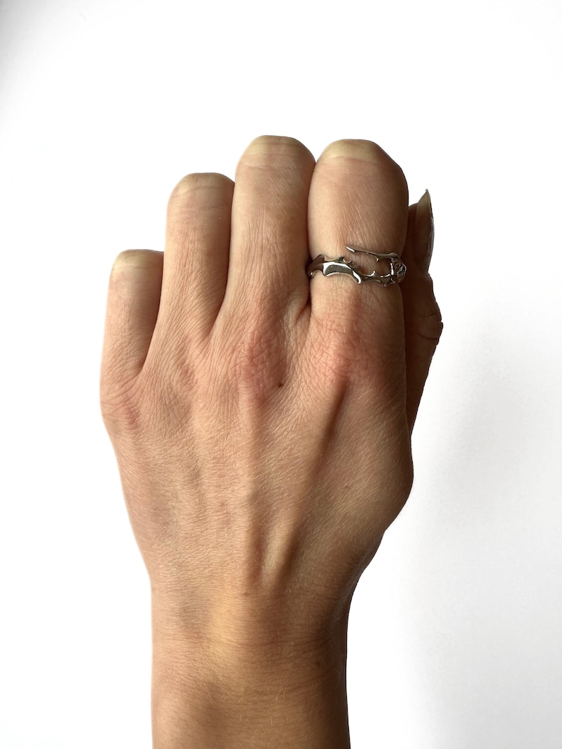 May include: A silver-colored ring with an organic, branch-like design. The ring is worn on a finger, showcasing its unique, nature-inspired aesthetic. The ring's open design adds to its modern appeal.