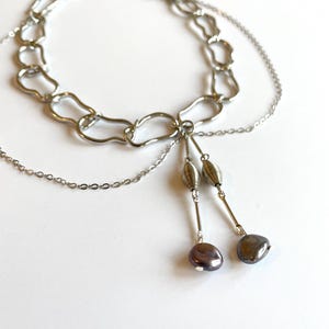 Op de afbeelding: Een zilverkleurige ketting met een chunky kettingontwerp. De ketting heeft een gelaagde uitstraling met een delicate ketting en twee bungelende strengen met decoratieve kralen en donkergrijs-paarse parels.