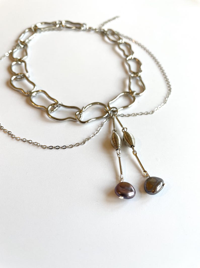 Op de afbeelding: Een zilverkleurige ketting met een chunky kettingontwerp. De ketting heeft een gelaagde uitstraling met een delicate ketting en twee bungelende strengen met decoratieve kralen en donkergrijs-paarse parels.