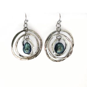 Può includere: Un paio di orecchini d'argento con un design circolare a strati. Ogni orecchino presenta tre anelli concentrici e una perla blu-verde scuro a forma di goccia sospesa al centro. Chiusura a gancio.